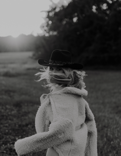 Girl running away from the camera but looking back, in black and white. She's wearing a black cowboy hat and a jacket